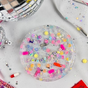 Puede incluir: Una bandeja redonda de resina transparente llena de pastillas de colores, signos de la paz y purpurina. La palabra "MEDICATED" está escrita con cuentas de letras. Una bola de discoteca y otros elementos decorativos están en el fondo.