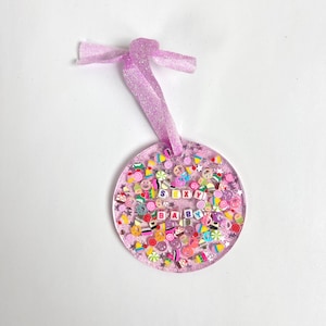 Pode incluir: Um enfeite redondo rosa com as palavras "SEXY BABY" escritas com contas de letras coloridas. O enfeite é preenchido com pequenas decorações coloridas em forma de doces e tem uma fita rosa brilhante para pendurar.