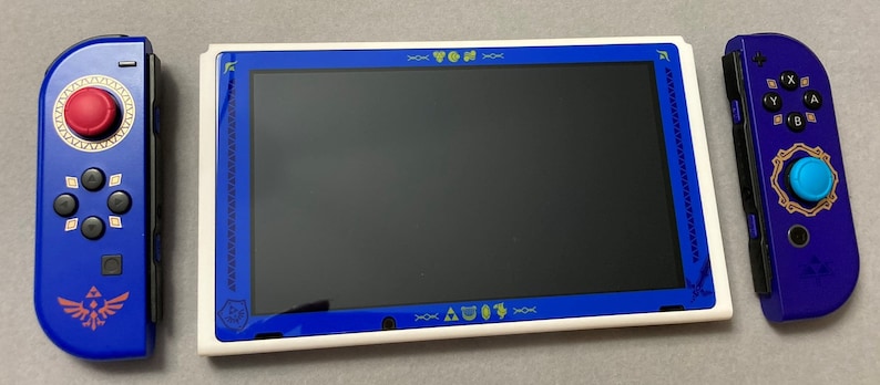 Puede incluir: Una consola Nintendo Switch blanca con un dise&ntilde;o tem&aacute;tico azul y dorado. La consola tiene una pantalla azul y un marco blanco. La consola est&aacute; acompa&ntilde;ada por dos mandos azules y morados con detalles dorados.