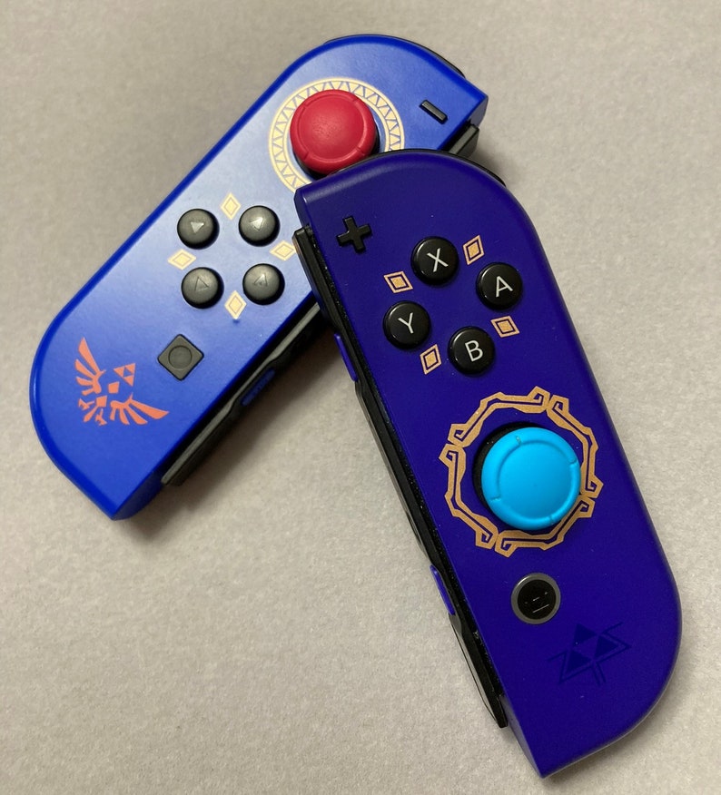 Puede incluir: Dos mandos Joy-Con para Nintendo Switch con un esquema de color azul y morado. El mando izquierdo tiene un bot&oacute;n rojo y un s&iacute;mbolo Triforce dorado. El mando derecho tiene un bot&oacute;n azul y un s&iacute;mbolo Triforce dorado.