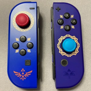 Puede incluir: Un par de mandos Joy-Con Nintendo Switch azules y morados con detalles dorados y un s&iacute;mbolo de Trifuerza. El Joy-Con izquierdo tiene un bot&oacute;n rojo y el Joy-Con derecho tiene un bot&oacute;n azul.