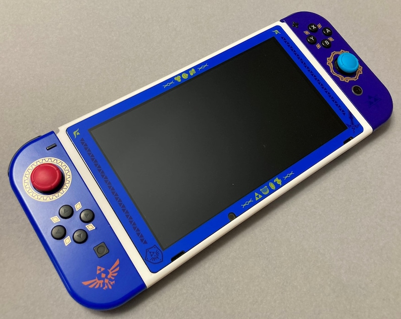 Puede incluir: Una consola Nintendo Switch azul y blanca con un dise&ntilde;o dorado y azul. La consola tiene una pantalla azul y un dise&ntilde;o dorado y azul en la parte posterior. La consola tiene un bot&oacute;n rojo en el lado izquierdo y un bot&oacute;n azul en el lado derecho.