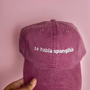 Puede incluir: Gorra de béisbol granate con la frase "se habla spanglish" bordada en blanco. La gorra está hecha de una tela suave y tiene una visera curva. El fondo es de color rosa liso.