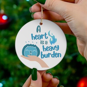 Op de afbeelding: Een witte kerstbal van keramiek met een blauwe en witte illustratie van een hart met vlammen en de tekst "A heart is a heavy burden".