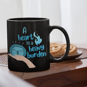 Op de afbeelding: Zwarte koffiemok met een blauwe en witte afbeelding van een hart met vlammen en de tekst "A heart is a heavy burden".