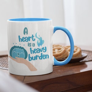 May include: White ceramic mug with a blue rim and handle. The mug features a blue and white illustration of a heart with flames and water droplets, and the text "A heart is a heavy burden".