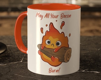 May include: White ceramic mug with an orange rim. The mug features a cartoon fire spirit holding a log with the text "May All Your Bacon Burn!"