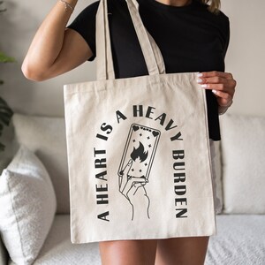Op de afbeelding: Een witte canvas tote bag met zwarte tekst die "A heart is a heavy burden" luidt. De tekst staat in een cirkel rond een zwart-witte illustratie van een hand die een tarotkaart met een vlam erop vasthoudt.