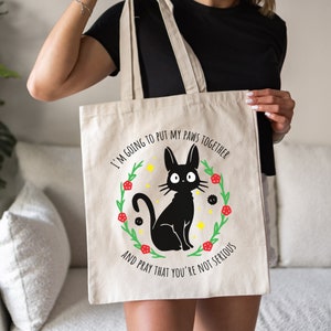 May include: A white canvas tote bag featuring a black cat illustration and the text "I'm going to put my paws together and pray that you're not serious".