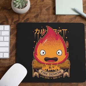 Może przedstawiać: Czarna podkładka pod mysz z kreskówkowym demonem ognia z uśmiechem. Demon jest pomarańczowy i czerwony z żółtym obramowaniem. Tekst "FIRE DEMON" znajduje się pod demonem, a tekst "ESTD. 2004" poniżej. Tekst "カルシファー" znajduje się nad demonem w języku japońskim.
