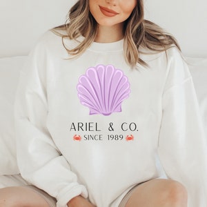 May include: White sweatshirt with a purple seashell graphic and the text "Ariel & Co. Since 1989" with two red crab illustrations.