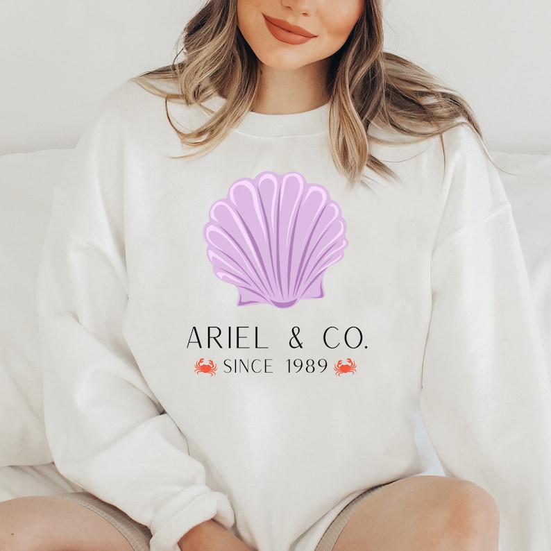 May include: White sweatshirt with a purple seashell graphic and the text "Ariel & Co. Since 1989" with two red crab illustrations.