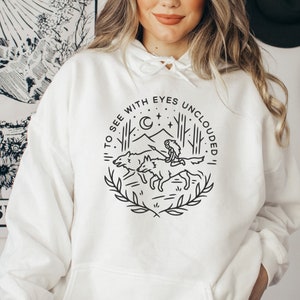 Puede incluir: Sudadera con capucha blanca con un diseño gráfico negro que representa a una mujer montando dos lobos en una escena de bosque. El texto "To see with eyes unclouded" está escrito en un círculo alrededor del gráfico.