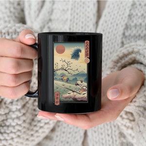 Puede incluir: Taza de cerámica negra con una ilustración de estilo japonés que representa un sol rojo, montañas y figuras. La obra de arte incluye texto japonés y una escena detallada con personajes y animales. La taza tiene un asa negra y un acabado brillante.