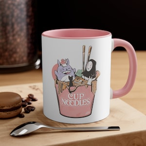 May include: White ceramic mug with a pink rim and handle. The mug features a cartoon illustration of Totoro and No Face from the movie Spirited Away, enjoying a bowl of instant noodles. The text "Cup Noodles" is printed on the bowl.