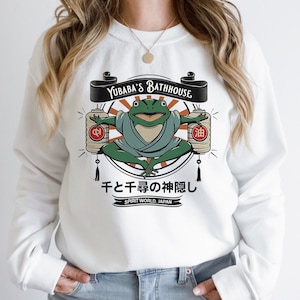 Op de afbeelding: Witte trui met een grafische print van een groene kikker die in meditatieve houding zit. De kikker draagt een blauwe robe en houdt twee rode lantaarns vast. De tekst "Yubaba's Bathhouse" en "千と千尋の神隠し" staat op de trui gedrukt.