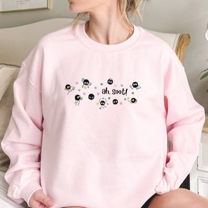 Op de afbeelding: Roze sweatshirt met een afbeelding van zwarte roetsprites uit de film 'Mijn buurman Totoro'. De afbeelding bevat de tekst 'ah, soot!' en sterren in verschillende kleuren.