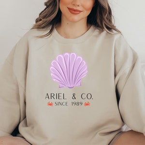 May include: A beige sweatshirt with a purple seashell graphic and the text "Ariel & Co. Since 1989" with two red crab illustrations.