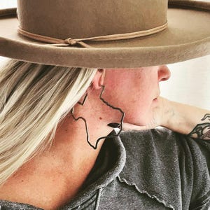 May include: A person wearing a brown hat and a grey shirt with a large black earring shaped like the state of Texas.