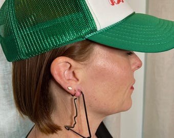 May include: A person wearing a green and white trucker hat with a red graphic. The person is wearing large black hoop earrings shaped like a race track.