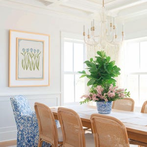 a dining room with a wooden table and chairs, a chandelier hanging above, and a framed artwork on the wall. The table is set with a vase of pink flowers, and the room has white walls and a window with white curtains.