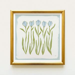 The image is a framed painting of a group of blue flowers with green stems, displayed on a white wall.