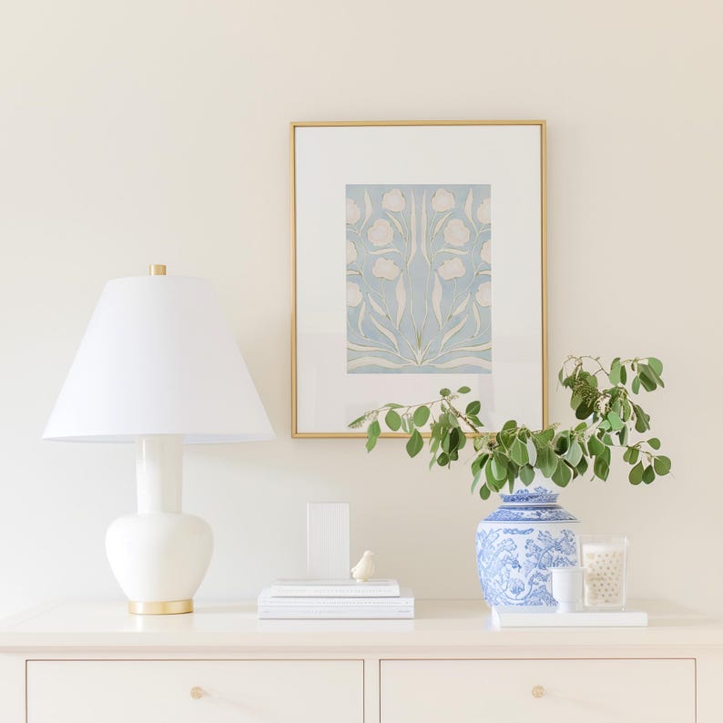a white dresser with a lamp, a framed picture, a blue and white vase with greenery, and some books and a small bird figurine on top.