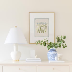 a white dresser with a lamp, a framed picture of flowers, a vase with greenery, and some books and decorative items on top.