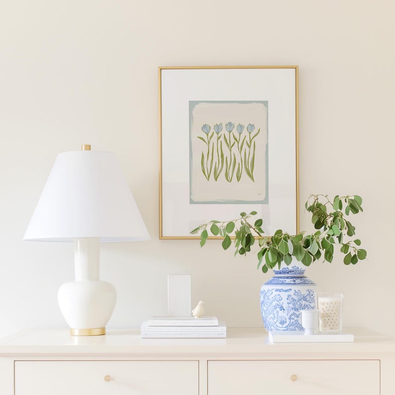 a white dresser with a lamp, a framed picture of flowers, a vase with greenery, and some books and decorative items on top.