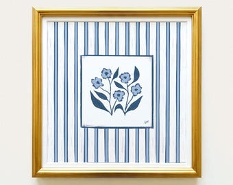 May include: A square framed artwork featuring a central white panel with a hand-painted illustration of blue flowers and dark blue leaves. The background consists of vertical light blue and white stripes. The piece is enclosed in a gold frame.