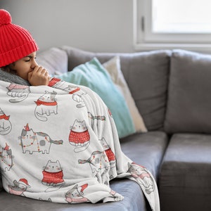 Könnte beinhalten: Eine Person trägt eine rote Strickmütze mit einem weißen Bommel und ist in eine weiße Fleece-Decke mit einem sich wiederholenden Muster aus grauen Katzen mit roten Schals und Weihnachtslichtern eingewickelt. Die Decke liegt auf einem grauen Sofa.