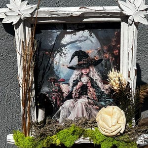May include: A framed artwork featuring a fantasy scene of a witch in a forest, holding a teacup. The artwork is in a distressed white wooden frame, decorated with dried plants and moss. The witch wears a black hat and gown.