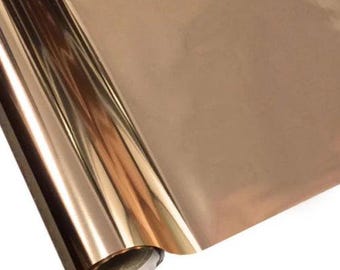 May include: A roll of metallic rose gold foil material with a smooth, reflective surface. The material is tightly wound around a cylindrical core, showing a high-gloss finish that catches the light, creating bright highlights and deep shadows.