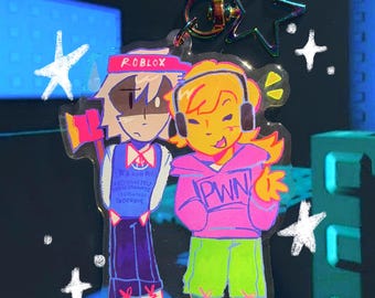 May include: A clear acrylic keychain featuring two cartoon characters. One character wears a Roblox hat and holds an axe, the other wears headphones and a pink hoodie with "PWN" on it. The keychain has a star-shaped clasp and white star accents.