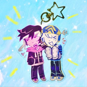 May include: A clear acrylic charm featuring two cartoon characters, one with black hair and a pink hoodie, the other with a blue and yellow helmet. A gold star-shaped keychain is attached at the top. The background is light blue with yellow streaks.