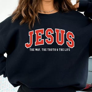 May include: Black sweatshirt with the word "JESUS" in red block letters with a white outline. Below the word "JESUS" is the text "THE WAY. THE TRUTH & THE LIFE" in white letters. The sweatshirt is a crew neck style.