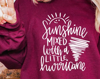 May include: Burgundy sweatshirt with white text that reads "Sunshine mixed with a little hurricane". The text is in a handwritten style with a sun and a tornado graphic.