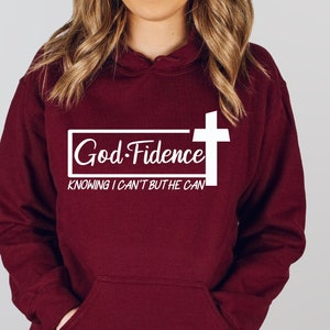 Może przedstawiać: Bordowa bluza z kapturem z białym nadrukiem, na kt&oacute;rym widnieje napis "God-Fidence" z krzyżem i tekstem "Knowing I can't but He can".