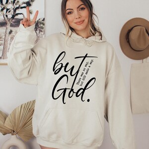 May include: Cream-colored hoodie with a front pocket and drawstring hood. The hoodie features the words "but God" in a black script font. Additional text reads "There was no way, but GOD made a way."