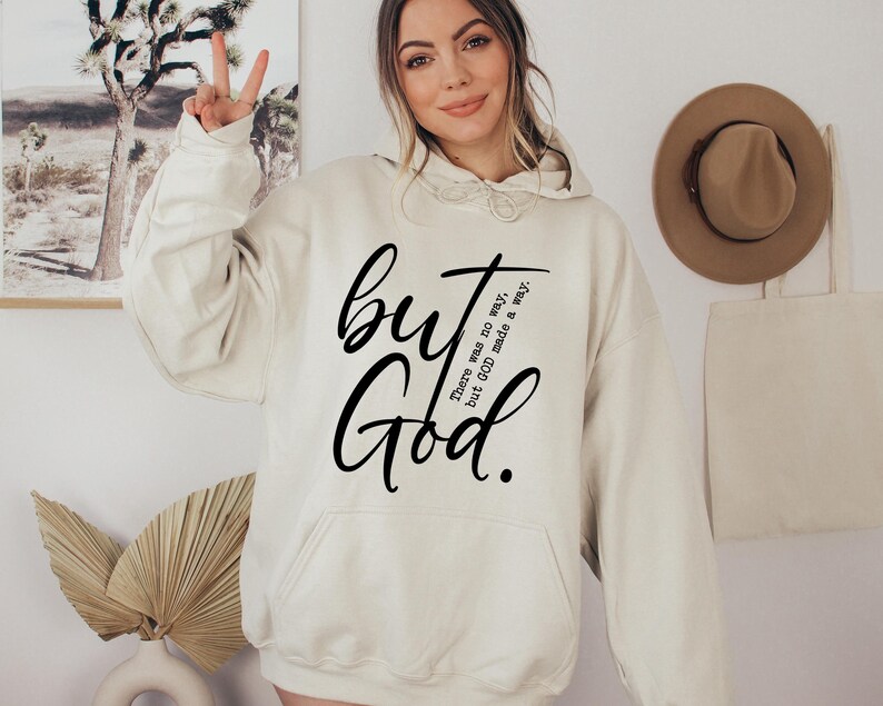 May include: Cream-colored hoodie with a front pocket and drawstring hood. The hoodie features the words "but God" in a black script font. Additional text reads "There was no way, but GOD made a way."