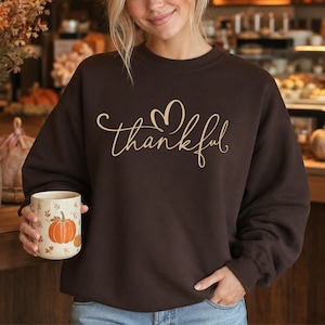 May include: A dark brown crewneck sweatshirt featuring the word thankful in a light gold cursive script with a heart-shaped flourish above the letter k. The sweatshirt is paired with a white mug decorated with orange pumpkin illustrations.