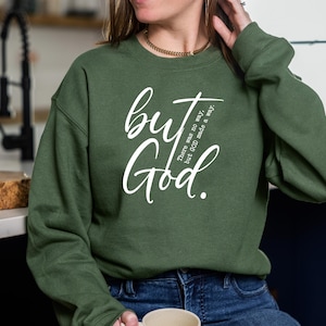May include: A dark green sweatshirt with the words "but God" in white script. The text "There was no way, but GOD made a way" is also printed on the shirt. The person is wearing blue jeans.