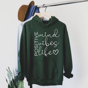 May include: A dark green hooded sweatshirt with the text "POSITIVE mind vibes life" in white lettering with small hearts.
