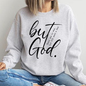 May include: A light gray sweatshirt with the words "but God" in large, black script. The phrase "There was no way, but GOD made a way" is printed in smaller, black text. The sweatshirt is a casual style.
