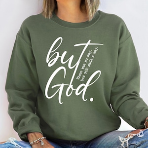 May include: Olive green sweatshirt with the words "but God" in a white script font. The text "There was no way, but GOD made a way" is also printed on the sweatshirt. The sweatshirt is a crew neck style.