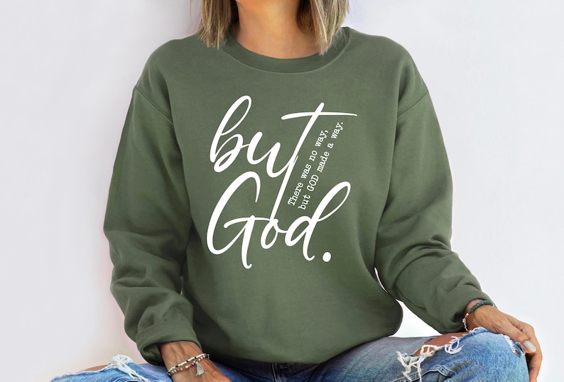 May include: Olive green sweatshirt with the words "but God" in a white script font. The text "There was no way, but GOD made a way" is also printed on the sweatshirt. The sweatshirt is a crew neck style.
