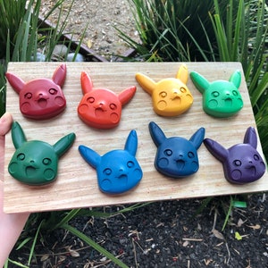 May include: Eight colorful crayon molds shaped like Pikachu, a popular cartoon character. The molds are arranged in a rainbow pattern, starting with red and ending with purple.