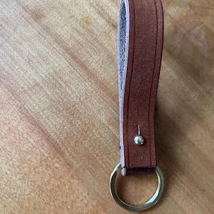 May include: A brown leather keychain with a gold-colored key ring. The leather loop is riveted and has a textured surface. The keychain is shown on a wooden surface.