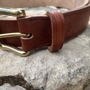 Puede incluir: Primer plano de un cinturón de cuero marrón con una hebilla dorada. El cinturón está hecho de cuero texturizado y presenta un diseño clásico. La hebilla es rectangular y brillante. El cinturón mide probablemente 3,18 cm de ancho.