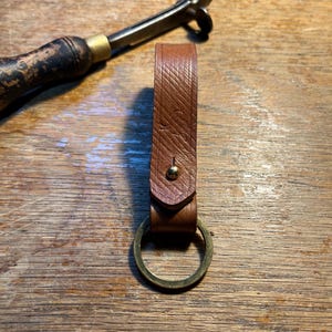 Può includere: Un portachiavi in pelle marrone con un portachiavi in ottone e un piccolo rivetto in ottone. Il cinturino in pelle è impresso con un motivo diagonale e le iniziali "S C". Il portachiavi è su una superficie di legno.